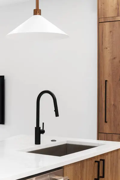 A kitchen faucet detail with wood cabinets, a black faucet, and gold light fixture hanging above the  marble countertops.