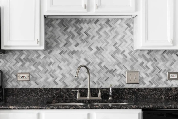 A kitchen faucet detail with white cabinets, a marble herringbone tile backsplash, and granite countertop.