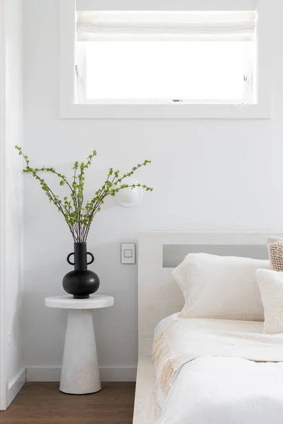 A bedroom detail with a neutral colored room and a tall plant sitting on a bedside table next to a short bed.