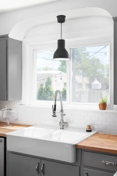 A kitchen sink detail with a butcher block wood countertop, white subway tile backsplash, grey cabinets, and a black light over a farmhouse sink.