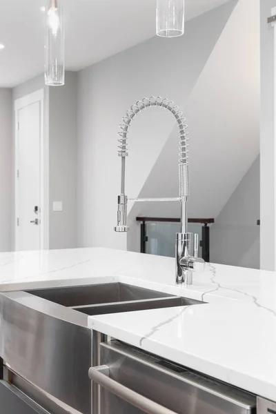 A kitchen faucet detail with a stainless steel apron sink, polished chrome faucet, and glass light fixtures hanging over the marble countertop island.