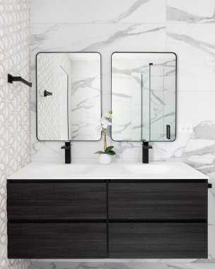 Yüzen ahşap bir dolabı, mermer döşeli duvarları, beyaz lavabosu ve siyah musluğu ve siyah çerçeveli aynaları olan bir banyo ayrıntısı..