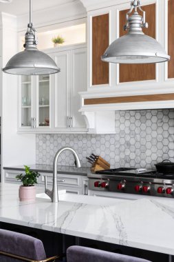 Büyük bir mermer adanın üzerinde asılı metal ışıklı bir mutfak müfrezesi, beyaz bir çiftlik evindeki paslanmaz musluk, ve beyaz dolaplarla süslü mermer bir altıgen..