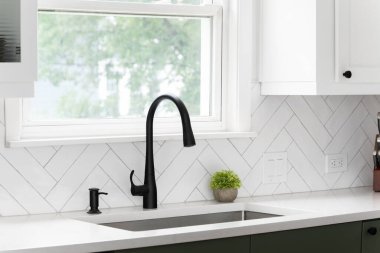 Lüks bir mutfakta beyaz ringa kemikli fayanslarla çekilen lavabo ayrıntıları. Beyaz mermer tezgah, beyaz ve yeşil dolaplar ve siyah bir musluk..