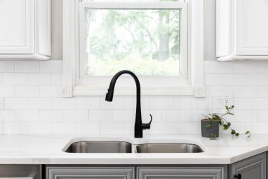 Mutfak lavabosu detayı, siyah musluk, beyaz metro fayansları, mermer tezgahın üzerine bitki, ve beyaz ve gri dolaplar..