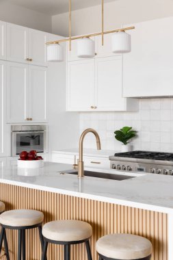 Beyaz dolapları ve kare döşemeleri olan bir mutfak detayı, bir şelale mermer tezgahı ve ahşap bölme paneli, ve altın bir aydınlatma armatürü, donanım ve musluk. Marka ya da etiket yok.
