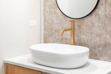 Banyoda altın musluğu olan bir lavabo ayrıntısı beyaz mermer tezgahta batan bir tekne ahşap dolap ve kahverengi desenli duvar kağıdında siyah bir ayna..