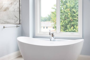 Lüks bir banyoda, krom musluğu, büyük pencereleri ve mermer kaplama duşu olan beyaz bir küvet detayı..