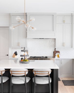 Açık gri dolapları, altın aksesuarları, beyaz mozaik döşemeleri ve büyük gri bir adada oturan bar tabureleri olan bir mutfak..