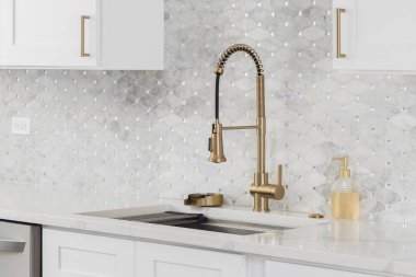 Altın musluğu olan bir mutfak, beyaz mermer mozaik döşeme, beyaz dolaplar ve beyaz bir tezgah..