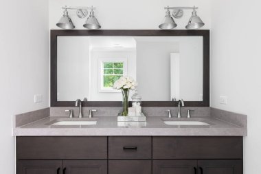 Ahşap bir dolabı ve çerçeveli aynası olan bir banyo, kahverengi kuvars tezgahı, parlak krom lambalar ve muslukları olan bir banyo..