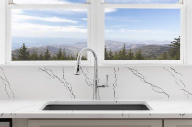 Koyu granit lavabosu, krom musluğu, beyaz mermer tezgahı ve büyük pencerelerden dağların ve mavi gökyüzünün manzarası olan bir mutfak detayı..