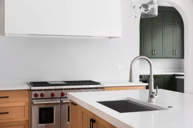 Beyaz mermer bir adanın tezgahında paslanmaz lavabo ve musluğu olan bir mutfak detayı paslanmaz çelik soba, ahşap dolaplar ve yeşil dolaplı bir kiler..