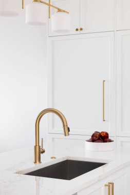 Beyaz bir mutfakta altın bir musluk mermer tezgah, paslanmaz çelik lavabo ve beyaz dolaplarda altın donanım..