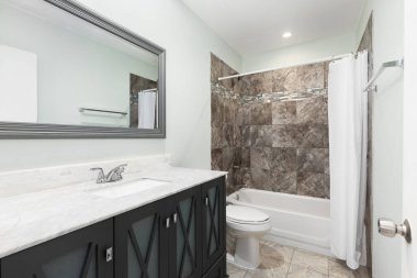 Buzlu cam kapıları, beyaz mermer tezgahı, döşemeli döşemesi ve kahverengi fayanslı duşu olan gri bir banyo..