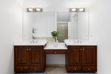 Ahşap dolaplı bir banyo, beyaz mermer tezgah, krom musluklar ve duşa bakan büyük bir ayna..