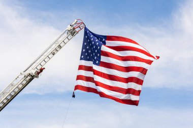 Rüzgarda dalgalanan bir Amerikan bayrağı. Arka planda mavi bir gökyüzü olan itfaiye merdiveninden sarkıyor. Amerikan bayramını kutluyor..