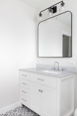 Beyaz dolabı, gri kuvars tezgahı, siyah ışık armatürü ve gri, beyaz ve siyah döşemesi olan bir banyo detayı..