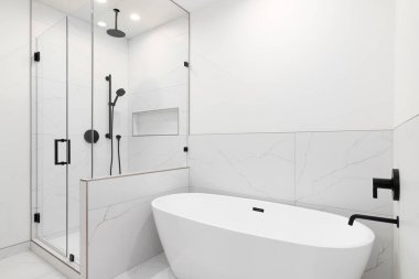 Banyoda beyaz bir standalon küvet ve beyaz mermer döşemeler ve siyah musluklarla banyo..