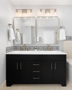 Siyah bir makyaj dolabı, bronz musluklar, aydınlatma armatürleri, döşeme döşemeleri ve beyaz tezgahı olan bir banyo detayı..