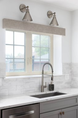 Mutfak musluğu, gri dolaplar, paslanmaz çelik musluk ve lavabo ve pencerenin üzerine monte edilmiş bronz ışıklar..