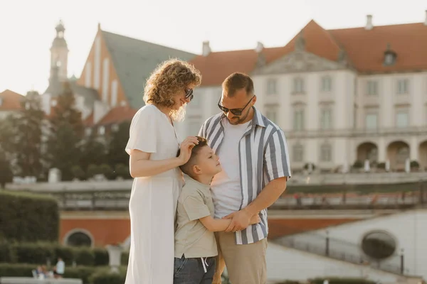 Father, mother and son are hugging in an old European town. Happy family in the evening. Mommy is stroking her little boys head in the background of the palace at sunset.