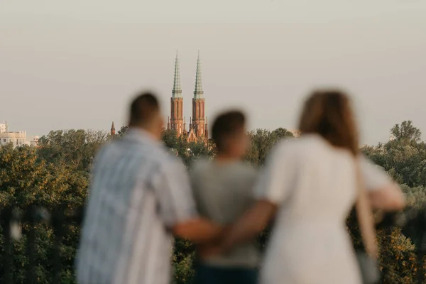 A photo from the back of father, mother, and son who are out of focus and staring at the old European church. The family of tourists is enjoying the town sight in the evening. Tourists at sunset.