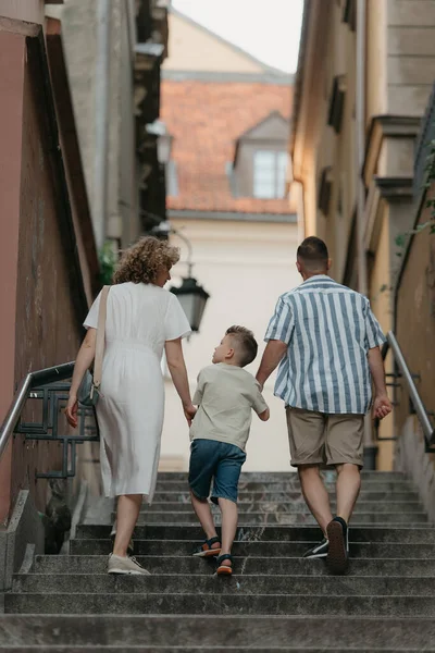 A full-length photo from the back of a family which is climbing stairs in an old European town. A happy father, mother, and son are holding hands and having fun in the evening.