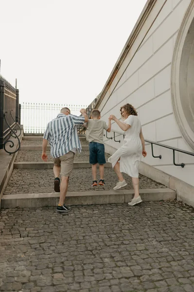 A photo from the back of a family is having fun while climbing the stairs of the palace in an old European town. A happy father, mother, and son are holding hands in the evening.