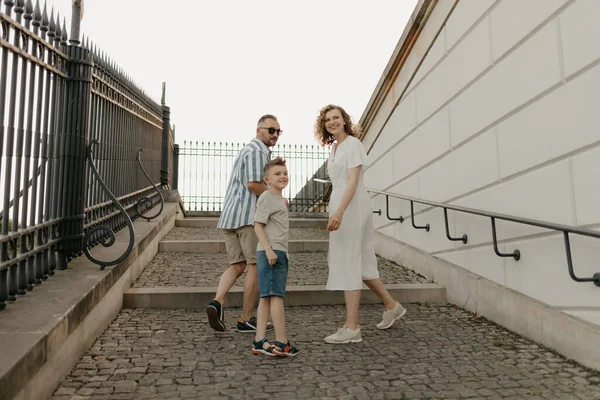 A photo from the back of a family is looking back while climbing the stairs of the palace in an old European town. A happy father, mother, and son are holding hands in the evening.