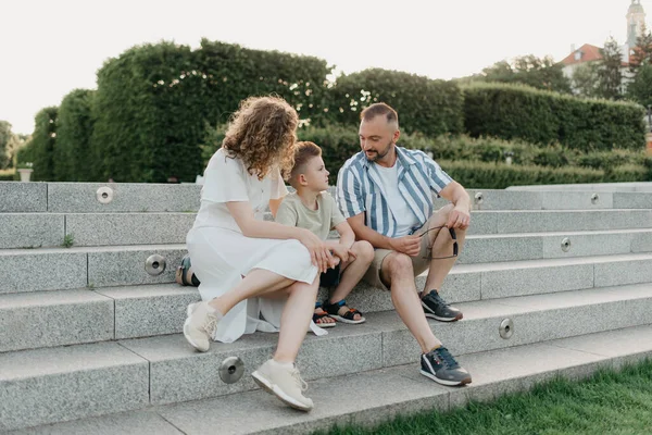 Father, mother, and son are sitting on the steps in the garden of an old European town. Dad is discussing important themes with his smiling family in the park in summer at sunset