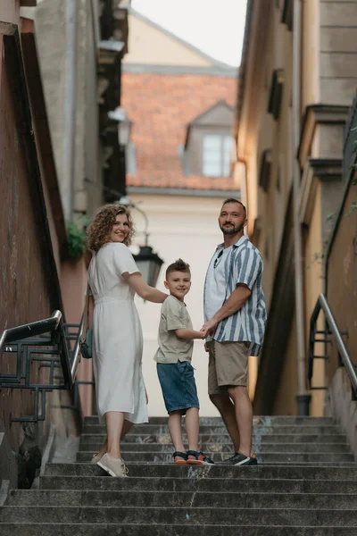 A full-length photo from the back of a family which is turning around while climbing stairs in an old European town. A happy father, mother, and son are holding hands and having fun in the evening.