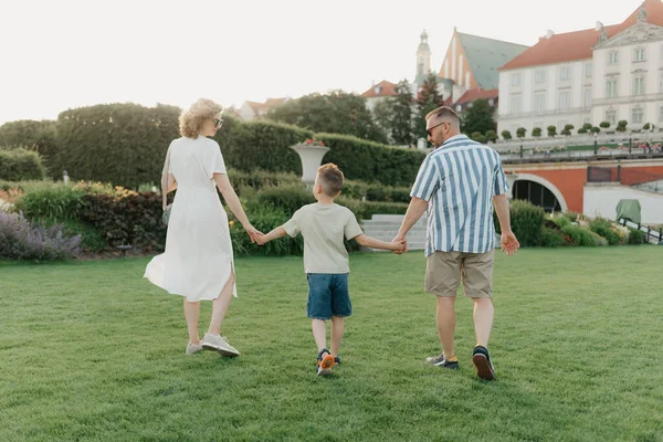 A photo from the back of a family is strolling the garden of the palace in an old European town. A happy father, mother, and son are holding hands and having fun at sunset