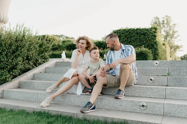 Father, mother, and son are sitting on the steps in the garden of an old European town. Happy family in the evening. Dad is discussing important themes with his family in the park at sunset.