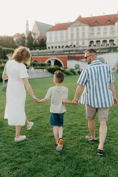 A photo from the back of a family is strolling the garden of the palace in an old European town. A happy father, mother, and son are holding hands at sunset.
