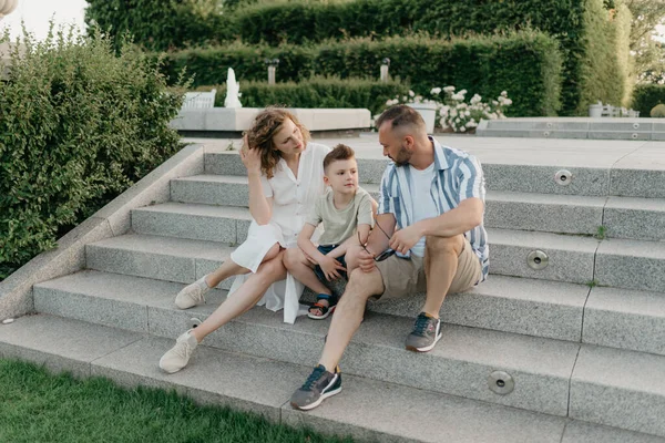 Father, mother, and son are sitting on the steps in the garden of an old European town. Happy family in the evening. Dad is talking with his family in the park at sunset.