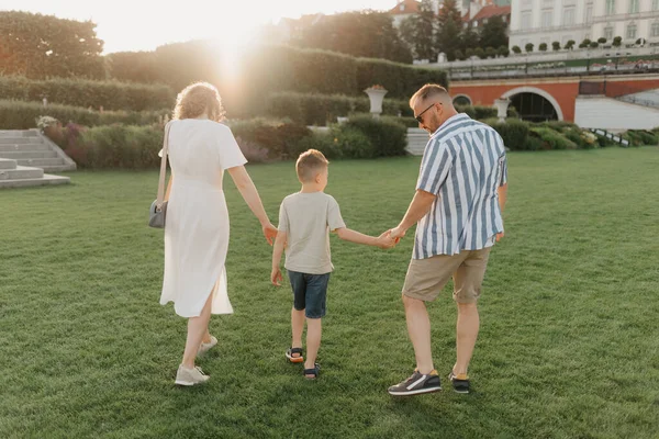 A photo from the back of a family is strolling the garden of the palace in an old European town. A happy father, mother, and son are holding hands in the evening.