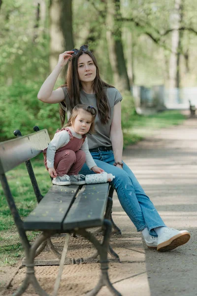 一位母亲在绿色公园的长椅上与她的小女儿靠得很近 一个穿着天鹅绒的女幼儿正在花园里和妈妈玩耍 图库图片