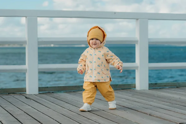 一个穿着黄色夹克和裤子的快乐的蹒跚学步的幼儿在码头上漫步 在波罗的海附近 一个头戴头巾的小女孩在玩得很开心 图库图片