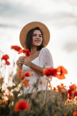 Kısa beyaz elbiseli ve saman şapkalı güzel bir kadın akşamları gelinciklerle birlikte tarlada ve elinde haşhaş, yaz kırsal doğa çiçekleri, kadın dinlendirici, kırsal basit hayat