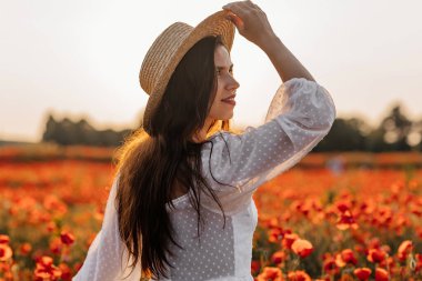 Kısa beyaz elbiseli ve saman şapkalı güzel bir kadın akşamları gelinciklerle birlikte tarlada ve elinde haşhaş, yaz kırsal doğa çiçekleri, kadın dinlendirici, kırsal basit hayat