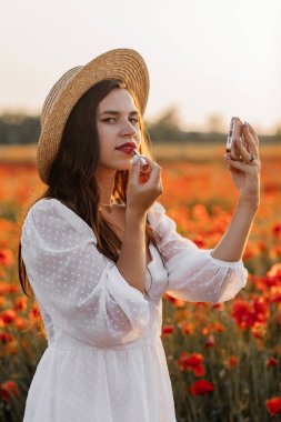 Kısa beyaz elbiseli ve saman şapkalı güzel bir kadın akşamları gelinciklerle birlikte tarlada ve elinde haşhaş, yaz kırsal doğa çiçekleri, kadın dinlendirici, kırsal basit hayat