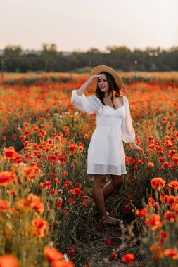 Kısa beyaz elbiseli ve saman şapkalı güzel bir kadın akşamları gelinciklerle birlikte tarlada ve elinde haşhaş, yaz kırsal doğa çiçekleri, kadın dinlendirici, kırsal basit hayat