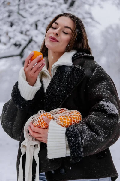 Winter holiday travel, Christmas day, New Year, beautiful happy woman portrait at snowy forest, nature woods, ski resort, leisure activity outdoors, Young Lady with mandarins