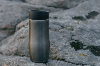 Dağların, taşların ve gün batımının ve bulutların yansıtıldığı bir nehrin arka planında turist termosu kupası. Seyahat, yürüyüş, spor, sağlıklı yaşam tarzı.