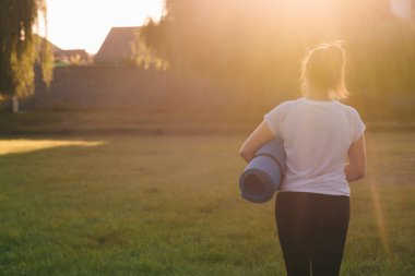 Arkadan bak. Spor kıyafetli bir kadın gün batımında yoga minderi tutuyor. Açık hava sporları.