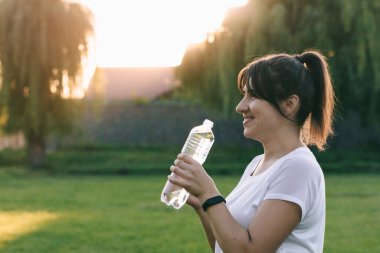 Şeffaf bir su şişesi tutarken neşeli sportif kız çekici bir şekilde gülüyor. Gün batımında egzersiz. Zindelik ya da yoga..