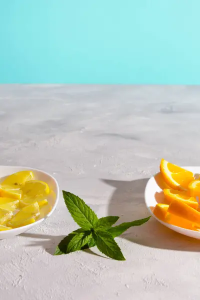 Beyaz tabakta limon ve portakal dilimlerinin, beyaz bir masanın üstünde nane filizi ile yakın plan fotoğrafı. Arka plan parlak bir turkuaz mavisi..