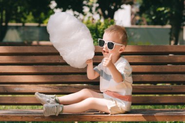 Küçük bir çocuk bankta oturuyor güneş gözlüğü takıyor ve pamuk şeker yiyor. Mutlu çocuk