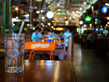 Bir bardak buzlu su, ahşap masa üzerinde saman keskin odak, bulanık, canlı bir restoran arka planı karşı ayarlı sıcak ışıklandırmalı.
